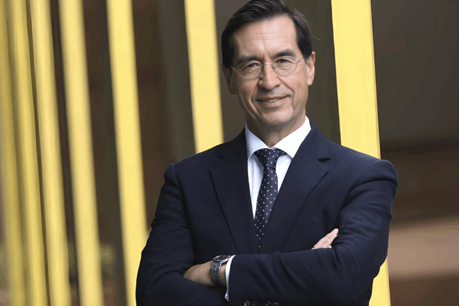 Mario Alonso Puig sonriendo, de brazos cruzados, vestido de traje formal frente a estructura arquitectónica amarilla.