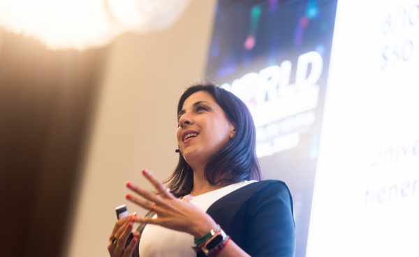 Fotografía de Gemma Ramírez hablando con entusiasmo en una conferencia internacional, con micrófono de diadema y proyector de fondo.