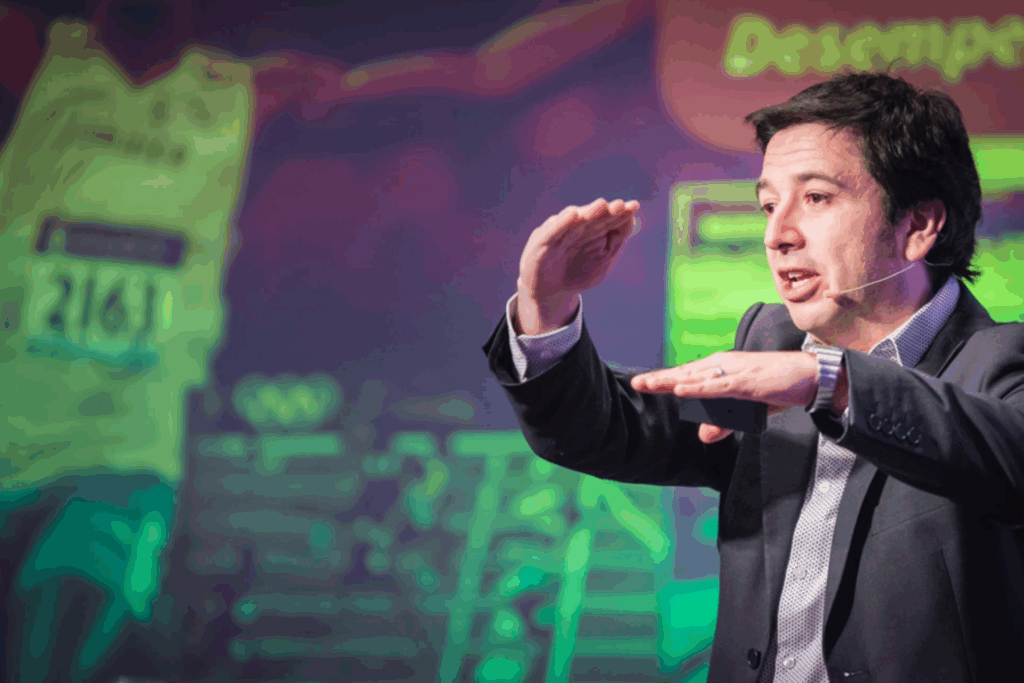 Fotografía de Rodrigo Saa durante una conferencia, expresando con gestos mientras utiliza un micrófono de diadema y un fondo colorido con elementos deportivos.