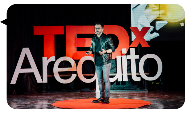 Ariel Benedetti dando una charla en el evento TEDx Arequito, vistiendo una chaqueta de cuero negra, camisa oscura y jeans, mientras sostiene un micrófono. En el fondo se observa el logo de TEDx con el nombre "Arequito" en letras grandes.