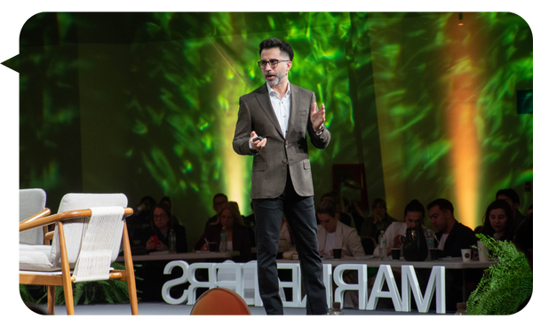 Ariel Benedetti dando una charla sobre marketing en un evento, vistiendo un saco marrón y jeans oscuros. Está de pie junto a una silla blanca en un escenario iluminado con luces verdes y amarillas. Al fondo, se ve al público atento.