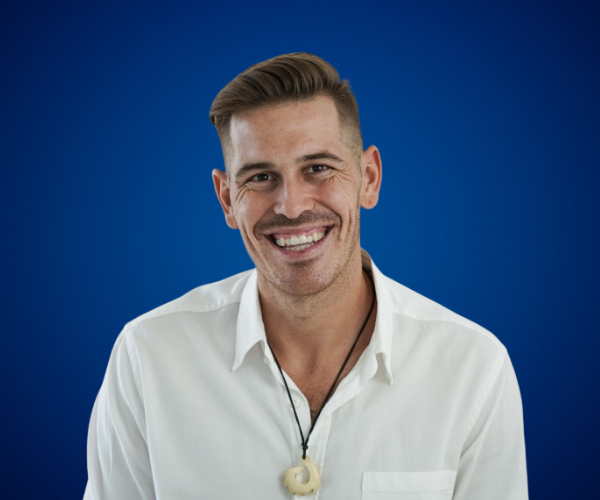 Fotografía de Ferran Tort sonriendo frente a un fondo azul sólido, vistiendo camisa blanca y collar artesanal con colgante en espiral.