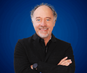 Fotografía de Luis Miguel Trujillo, conferencista internacional, sonriendo con los brazos cruzados sobre un fondo azul liso, vistiendo saco oscuro con camisa negra, en una imagen profesional de estudio.