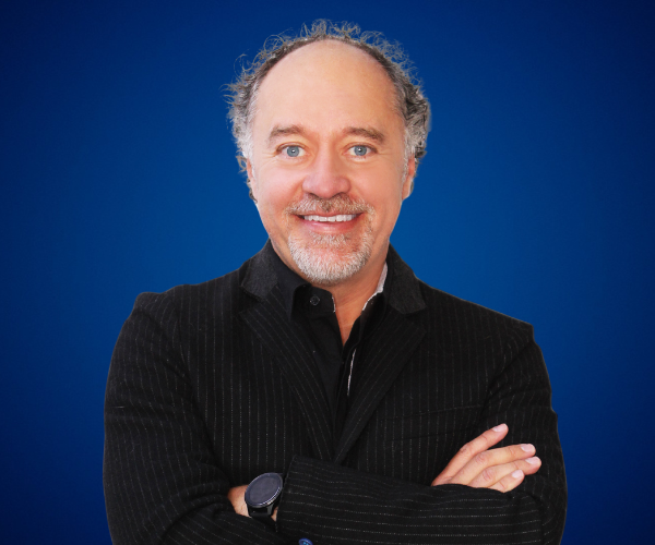 Fotografía de Luis Miguel Trujillo, conferencista internacional, sonriendo con los brazos cruzados sobre un fondo azul liso, vistiendo saco oscuro con camisa negra, en una imagen profesional de estudio.