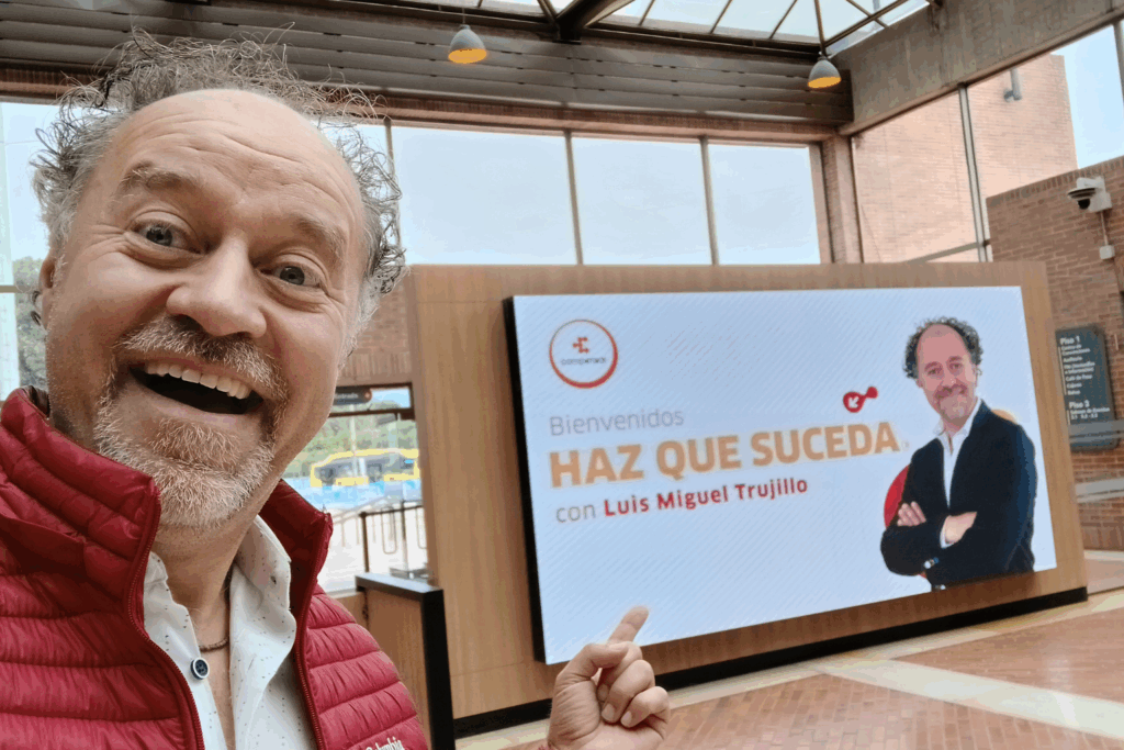 Fotografía de Luis Miguel Trujillo sonriendo y señalando una pantalla digital con el anuncio de su conferencia “Haz que suceda” en un espacio interior moderno y bien iluminado.