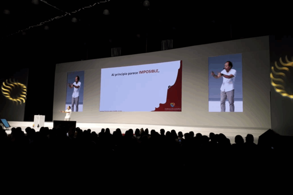 Fotografía de Luis Miguel Trujillo en una conferencia multitudinaria, en un escenario iluminado con pantallas gigantes que proyectan su imagen y una diapositiva con la frase “Al principio parece IMPOSIBLE”.
