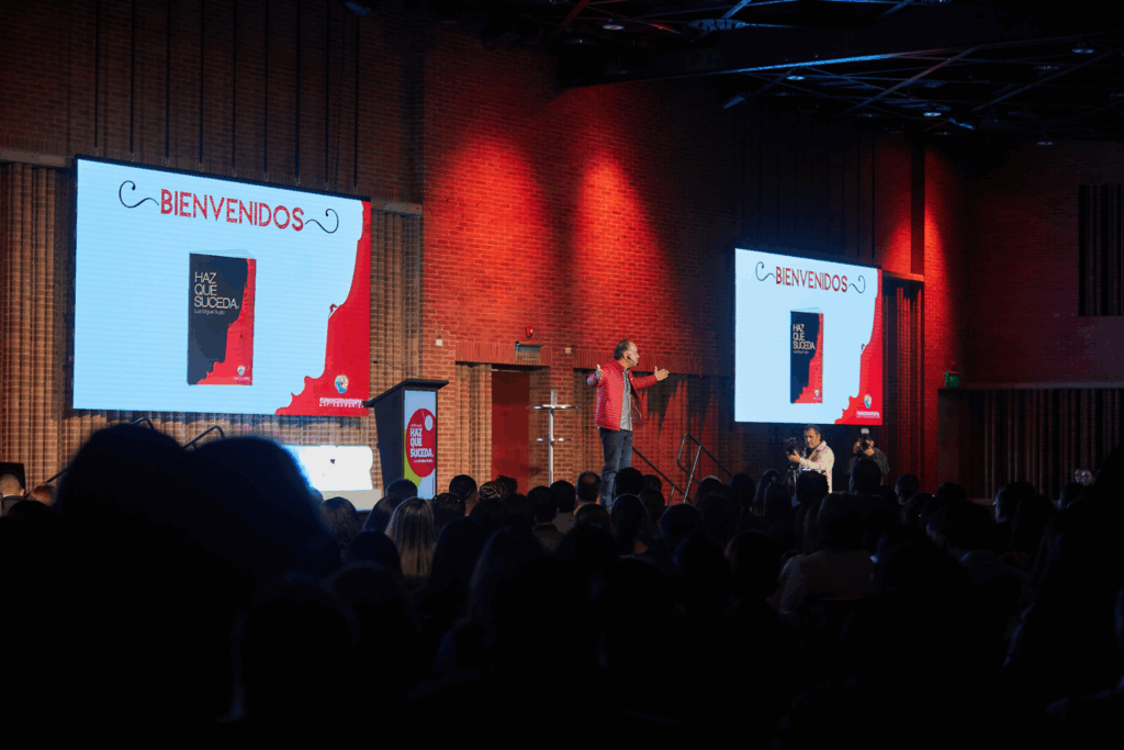 Fotografía de Luis Miguel Trujillo en tarima durante una conferencia, con pantallas que muestran la portada de su libro “Haz que suceda” y el mensaje de bienvenida, en un auditorio con iluminación cálida y paredes de ladrillo.