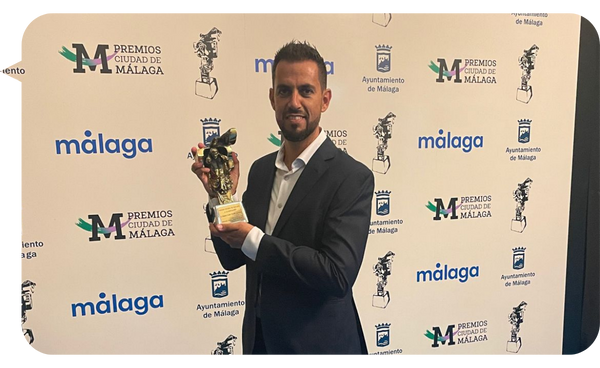 Miguel Ángel Ronda sosteniendo el Premio Ciudad de Málaga, destacando su contribución a la innovación educativa y tecnológica, con un fondo de logos de Málaga y el Ayuntamiento de Málaga.