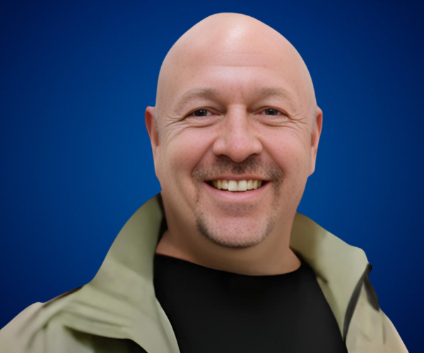 Fotografía de Pablo Nachtigall, conferencista calvo con barba de candado, sonriendo, vestido con camiseta negra y chaqueta beige, sobre un fondo azul liso.
