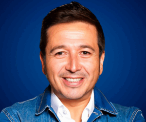 Fotografía de Rodrigo Saa, conferencista internacional, con cabello canoso, barba ligera y expresión serena, sobre fondo azul profesional, vestido con camisa blanca.