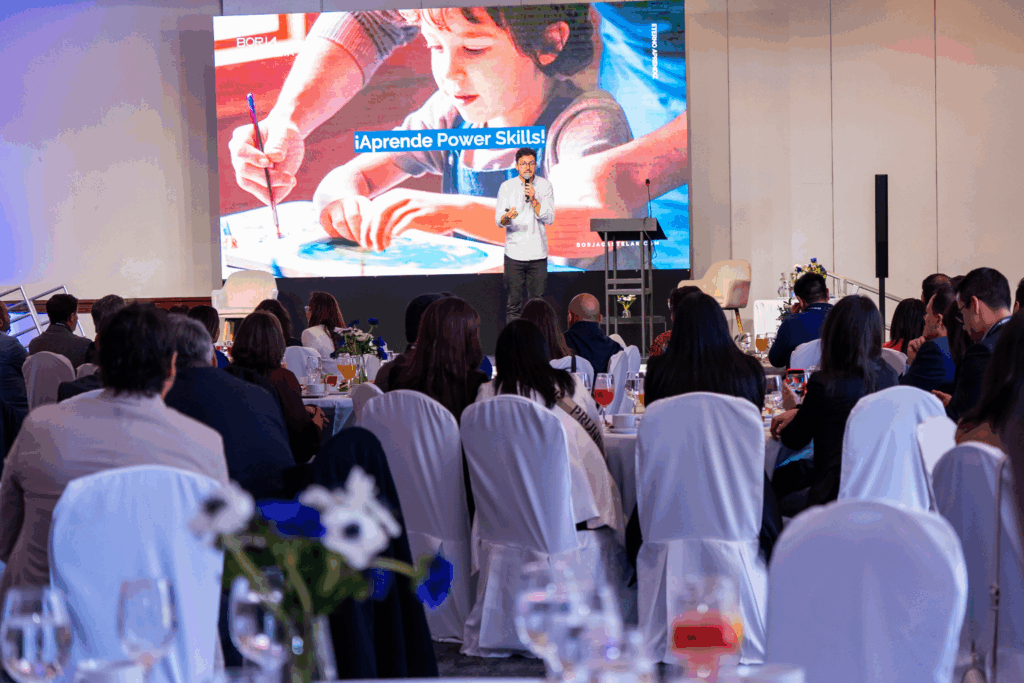 Fotografía de Borja Castelar dando una conferencia en un auditorio con público sentado en mesas redondas; en el escenario, una pantalla muestra el mensaje “¡Aprende Power Skills!”.