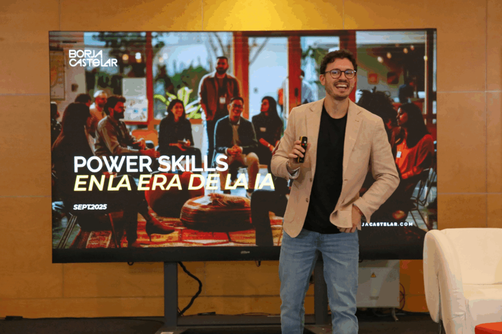 Fotografía de Borja Castelar durante una conferencia, sonriendo con micrófono en mano frente a una pantalla que dice “Power Skills en la era de la IA”, en un ambiente moderno y acogedor.