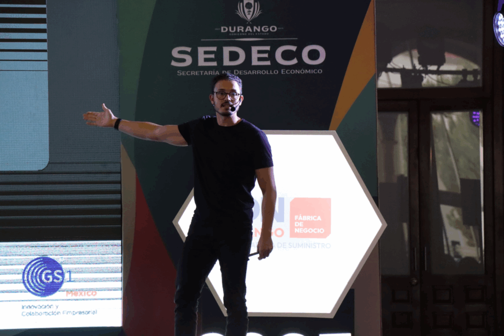 Fotografía de Borja Castelar en el escenario de un evento organizado por SEDECO Durango, usando micrófono de diadema y vestido de negro, señalando con el brazo extendido hacia una pantalla lateral.