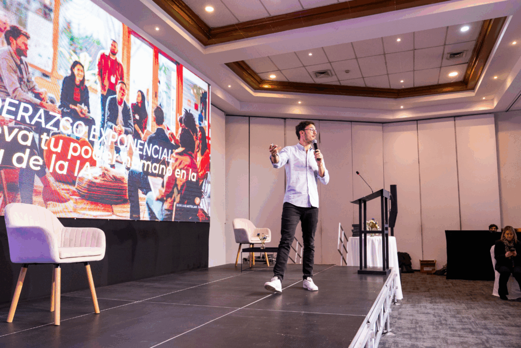 Fotografía de Borja Castelar ofreciendo una conferencia en un auditorio moderno, de pie sobre el escenario con micrófono en mano, frente a una pantalla que proyecta el mensaje “Liderazgo Exponencial”.