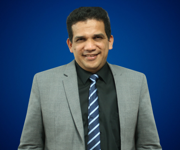 Fotografía de Jaime Montenegro, con fondo azul sólido, vistiendo traje gris, camisa negra y corbata a rayas, mostrando una expresión amable y profesional.