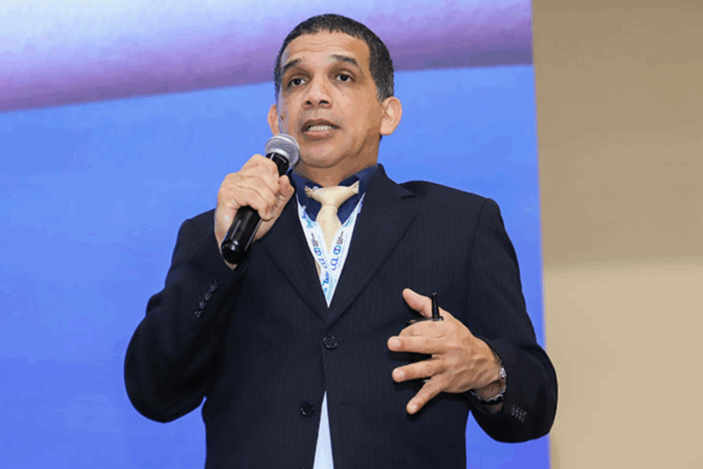 Fotografía de Jaime Montenegro durante una conferencia, vestido con traje oscuro y corbata clara, hablando al micrófono con fondo azul, portando credencial de evento tecnológico.