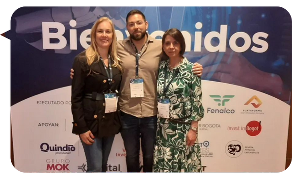 Nina Valderrama en un evento junto a dos colegas, frente a un cartel de bienvenida con logos de patrocinadores.