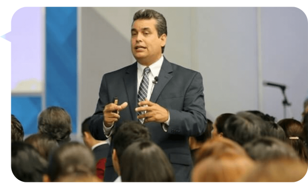 William Rodríguez dando una conferencia frente a un público, vestido con un traje oscuro y corbata.