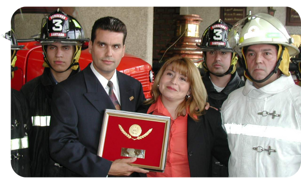 William Rodríguez recibiendo un reconocimiento, rodeado de bomberos, junto a una mujer.