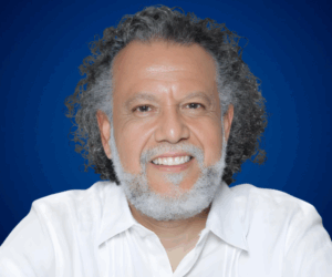 Fotografía de Alberto Linero, conferencista internacional, con camisa blanca y fondo azul profesional, mirando a cámara con una sonrisa cercana.