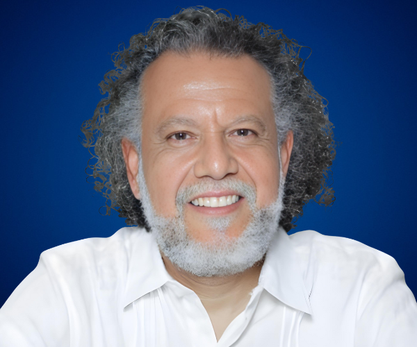 Fotografía de Alberto Linero, conferencista internacional, con camisa blanca y fondo azul profesional, mirando a cámara con una sonrisa cercana.
