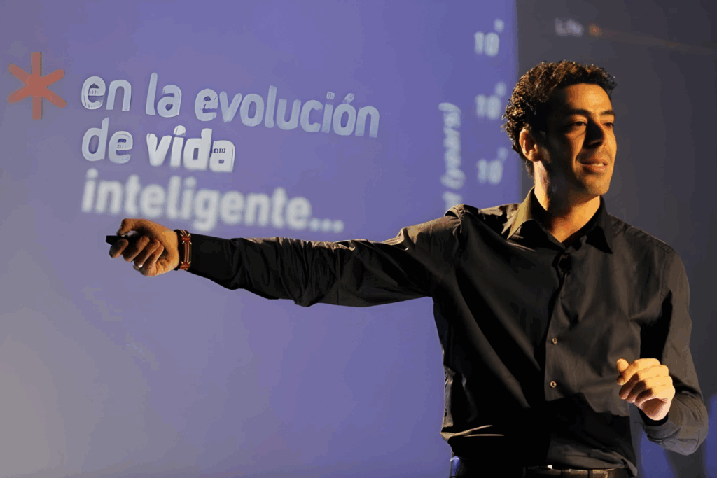 Fotografía de Gabriel Gurovich impartiendo una conferencia, señalando una presentación proyectada con la frase “en la evolución de vida inteligente...”, vistiendo camisa negra.