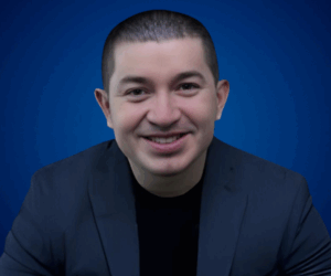 Fotografía de Gustavo Henao sonriendo frente a un fondo azul, con chaqueta oscura y camiseta negra, en un retrato profesional.