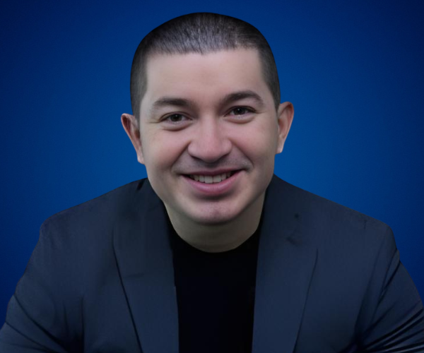 Fotografía de Gustavo Henao sonriendo frente a un fondo azul, con chaqueta oscura y camiseta negra, en un retrato profesional.
