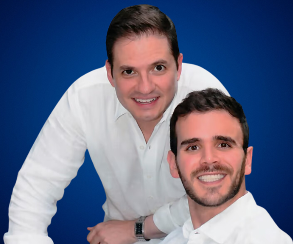 Kike y Gino sonriendo, ambos con camisas blancas, sobre un fondo azul.