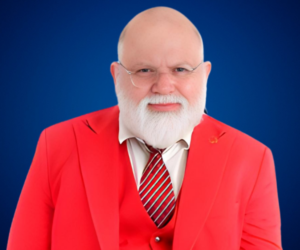 Hombre de edad madura, calvo y con barba blanca, vistiendo un traje rojo con chaleco y corbata a rayas rojas y blancas. Usa lentes y tiene una expresión amable. El fondo es azul sólido.