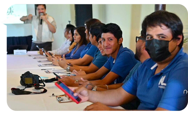 Participantes de un taller, vestidos con camisas azules, sentados en una mesa con dispositivos móviles en mano, mientras Ness Carballo, conferencista, les habla desde el frente de la sala.