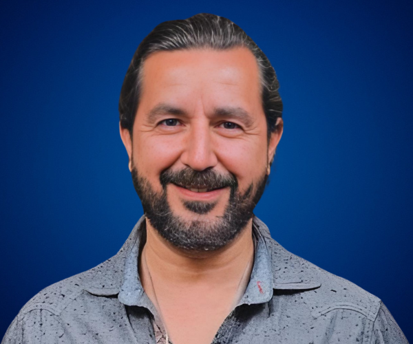 Fotografía de Daniel Villarreal, sonriendo con una camisa gris clara sobre un fondo azul, proyectando seguridad y cercanía.
