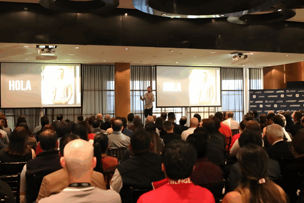 Fotografía de Daniel Villarreal dando una conferencia en un auditorio lleno, acompañado de pantallas con la palabra “HOLA” como introducción a su charla.