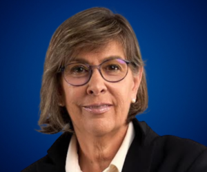 Retrato de Carmen Álvarez Basso, una mujer con gafas y cabello corto, vestida de manera profesional, con un fondo azul.