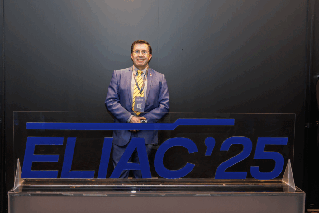 Fotografía de Rodrigo Rivera en el evento ELIAC’25, vistiendo traje azul con corbata amarilla y gafas, posando sonriente detrás del logotipo oficial del congreso.