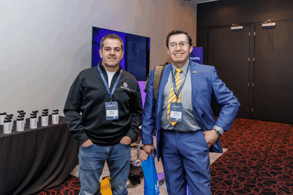 Fotografía de Rodrigo Rivera junto a otro participante en el evento ELIAC’25, vistiendo traje azul con corbata amarilla y gafas, posando en un entorno de congreso tecnológico.