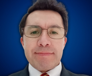 Fotografía de Rodrigo Rivera, con expresión serena, usando gafas, traje formal y corbata a rayas rojas y doradas, sobre un fondo azul profesional.