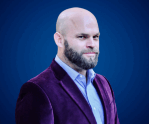 Alejandro Lanuque con saco de terciopelo violeta y camisa celeste, mirando con confianza sobre fondo azul.