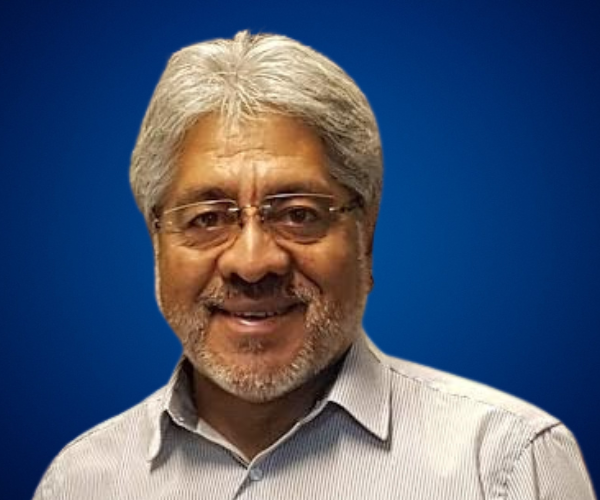 Carlos Ñanco, experto en recursos humanos, ventas y servicio al cliente, con cabello canoso y barba, sonriendo y vistiendo una camisa a rayas clara sobre un fondo azul.