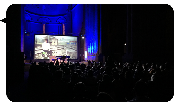 Antoni Tolmos en una presentación audiovisual en un auditorio oscuro, con una pantalla proyectando su imagen y el escenario iluminado con luces azules.