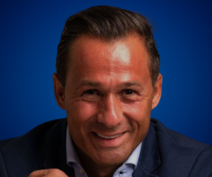 Retrato de Antonio Jiménez, experto en coaching y desarrollo del potencial humano, sonriendo frente a un fondo azul.