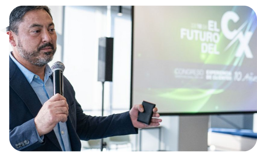 Claudio López Moreno hablando frente a una pantalla con el tema "El Futuro del CX," sosteniendo un micrófono y un control remoto, en un evento corporativo.