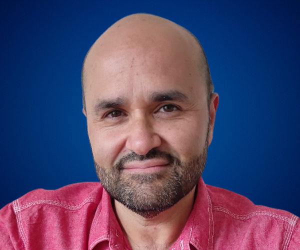 Fotografía de Fernando Veliz Montero, un hombre con barba y una camisa roja, sonriendo frente a un fondo azul. Fernando es un conferencista y consultor especializado en comunicación organizacional, resiliencia y liderazgo.