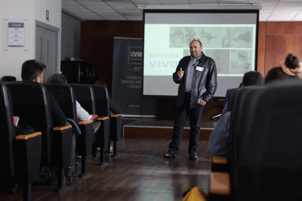 Fotografía de Fernando Veliz dando una conferencia en un auditorio universitario, con proyección sobre “Sistema Vivo” al fondo.