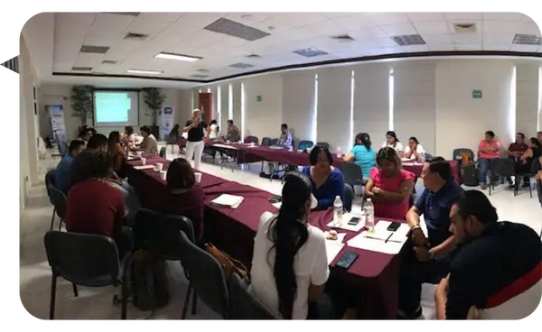 Mónica Reta, conferencista y coach, lidera una sesión de capacitación en estrategias de liderazgo en una sala de conferencias. Los asistentes están sentados en mesas en forma de U, participando activamente en discusiones grupales.