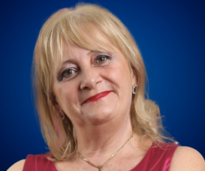 Mónica Reta, conferencista internacional y coach de vida, con fondo azul y vestido rosa, sonriendo a la cámara.