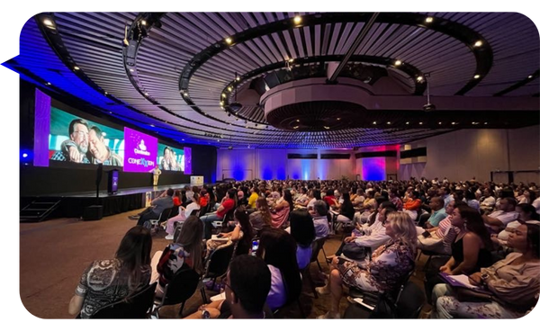 Conferencia llena de asistentes en un gran salón con un escenario iluminado y pantallas proyectando imágenes.