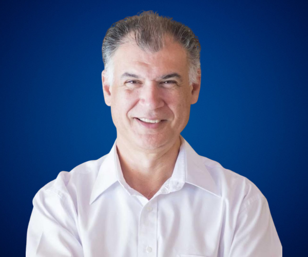 Retrato de Roberto Ravagnani, conferencista internacional experto en voluntariado, sostenibilidad y ESG, sonriendo con una camisa blanca frente a un fondo azul.