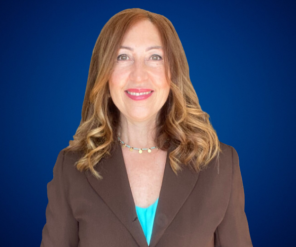 Retrato profesional de Sandra Cavallo sonriente con cabello largo y ondulado, vistiendo un blazer marrón sobre una blusa turquesa, con un collar delicado, sobre un fondo azul.