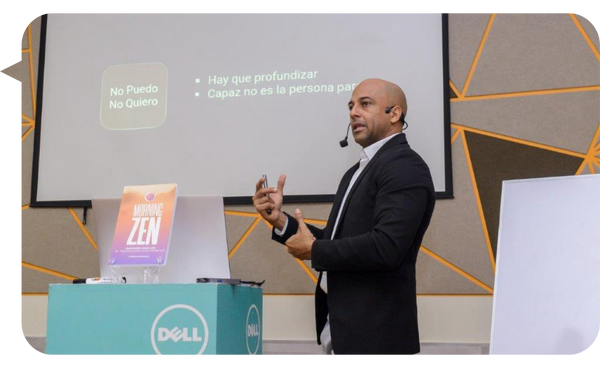 Adrián González dando una conferencia, con un micrófono de diadema, frente a una pantalla que muestra el texto "No Puedo, No Quiero" y un libro titulado "Morning Zen" a su lado.